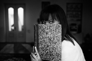 Pretty woman hides behind the book sitting at her living room. Monochrome photo with dust and grain...