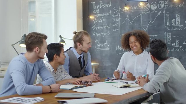 Group of multiethnic businesspeople sitting at desk in modern office and discussing work together, business charts are drawn on chalkboard in the background