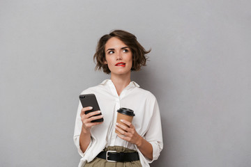 Photo of attractive woman 20s holding takeaway coffee and using mobile phone, isolated over gray...