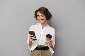 Photo of european woman 20s holding takeaway coffee and using cell phone, isolated over gray background