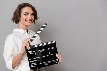 Photo of european woman 20s smiling and holding black clapperboard, isolated over gray background
