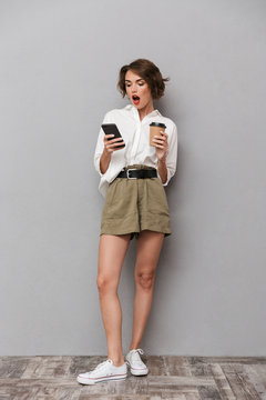 Full length image of excited european woman 20s holding takeaway coffee and using smartphone, isolated over gray background