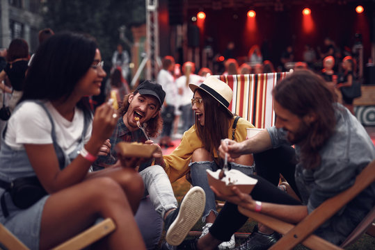 Portrait of cheerful hipster girl in hat feeding boyfriend with fork while sitting on folding chair. Blurred bearded man and girl in glasses eating and smiling