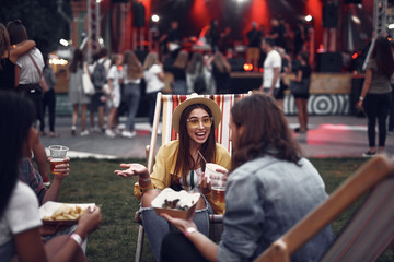 Obraz premium Portrait of cheerful girl in hat sitting on folding chair and chatting with young man. Stage and crowd on blurred background