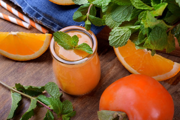 Orange smoothie in a glass jar surrounded by persimmon, pieces of oranges and mint against the...