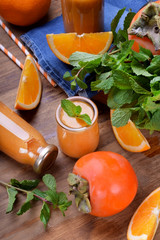 Orange smoothie in a glass jar surrounded by persimmon, pieces of oranges and mint against the wooden background