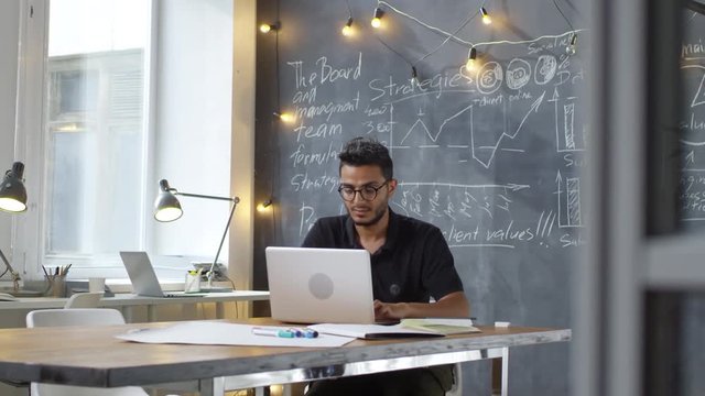 Tracking shot of focused young Asian man in glasses working on laptop computer while sitting at desk in modern office with business charts drawn on chalkboard