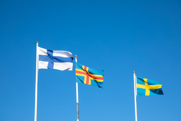 The flags of Finland, Åland Islands, and Sweden flying in the breeze against the backdrop of a clear blue sky; at Kökar, a municipality of the Åland Islands, Finland, in the Baltic Sea.