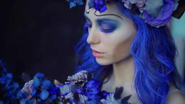 amazing halloween makeup and hairstyle in blue colors on young woman, girl is looking on flowers