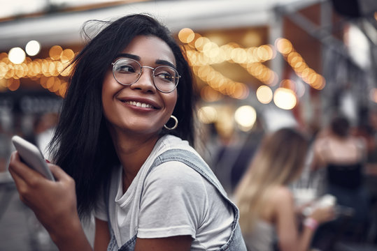 Portrait of hipster young lady in glasses holding smartphone and looking away with smile. Street with people on blurred background with bokeh effect