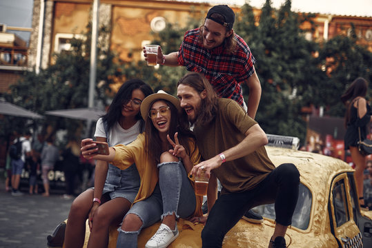 Portrait of stylish young people sitting on yellow car and taking picture with smartphone. Smiling bearded guys holding cups with cold beer while girl in hat showing victory sign