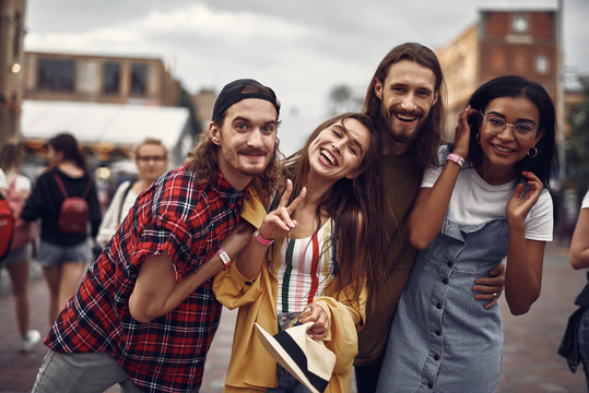 Portrait of cheerful hipster friends hugging and looking at camera with smiles. Lovely girl holding hat and showing victory gesture