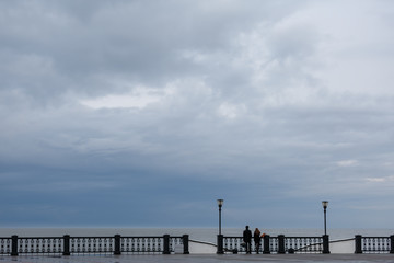 Young man and young woman stand around the openwork fence and look at the sea on a rainy day