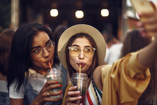 Portrait of young beautiful lady in hat taking photo with charming friend. Women drinking cold mint beverages