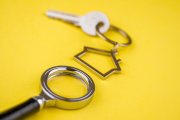 The concept of checking the legal purity of a private house or apartment. Metal keyring in the shape of a house with a metal key under a magnifying glass on a yellow background. Close up.