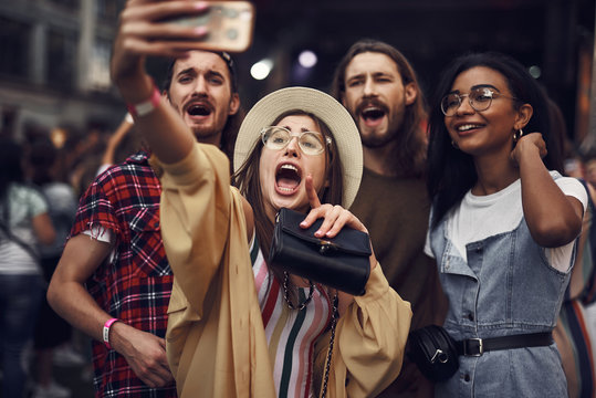 Waist up portrait of young lady in hat taking photo with smartphone while holding purse. Group of hipsters looking at camera and screaming with joy