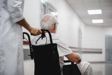 Back view portrait of old gentleman sitting in wheelchair while female medic standing behind
