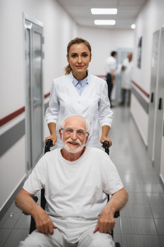 Vertical portrait of joyful bearded gentleman sitting in wheelchair while female medic standing behind and looking at camera with smile. Medical workers on blurred background