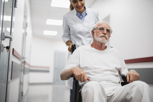 Portrait of serious bearded gentleman sitting in wheelchair while female medic looking at him with smile