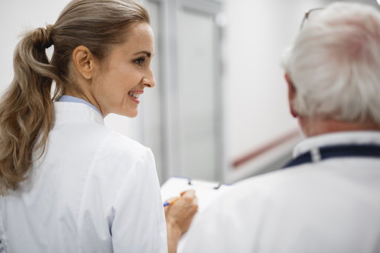 Back view portrait of beautiful young lady in white lab coat holding clipboard and chatting with old therapist. They heading to patient room