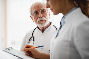 Portrait of bearded surgeon in glasses staring at young lady in white lab coat while she writing on clipboard. Focus on elderly man