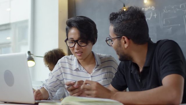 Handheld medium shot of two positive young people sitting at desk in modern office and discussing work, man helping woman with laptop computer