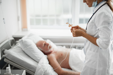 Cropped side view portrait of young lady in white lab coat checking IV infusion. Old man lying in hospital bed on blurred background