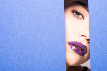 Face of young beautiful woman with a bright make-up and violet lips looks through a hole in violet paper.