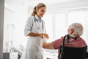 Portrait of young lady in white lab coat taking care of old man after surgery. She touching his arm and looking at him with smile