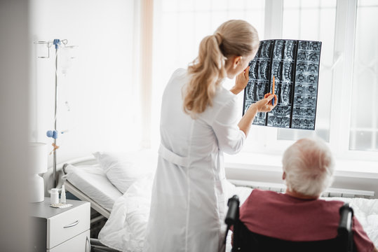 Back view portrait of young lady in white lab coat holding x-ray image and pointing at it while gentleman sitting in wheelchair