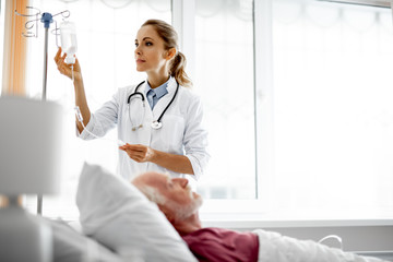 Waist up portrait of young woman in white lab coat checking intravenous drip in hospital room....