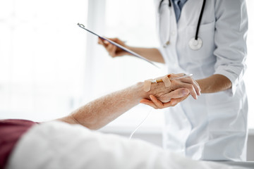 Cropped portrait of young lady in white lab coat holding clipboard and checking patient condition while he lying in bad. Focus on male arm