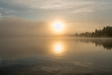 Beautiful golden Sun glows through fog at lake during early sunrise, summer, sky reflection in calm water