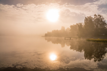 Foggy sunset with forest at long coast line, golden calm lake, Swedish countryside