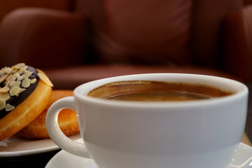 One white cup of hot coffee with light smoke, blurred doughnuts and armchair in background 