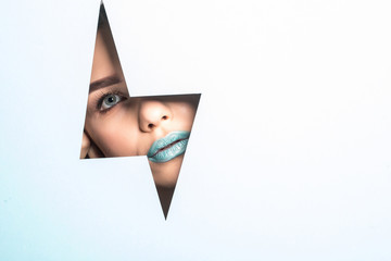 Face of a young beautiful girl with a bright make-up and puffy blue lips peers into a hole in blue...