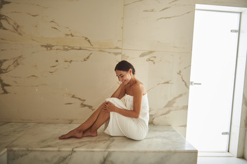 Happy hammam visitor wearing white towel and smiling while sitting on the marble surface - obrazy, fototapety, plakaty