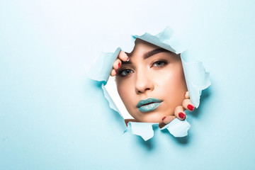Face of a young beautiful girl with a bright make-up and puffy blue lips peers into a hole in blue paper.