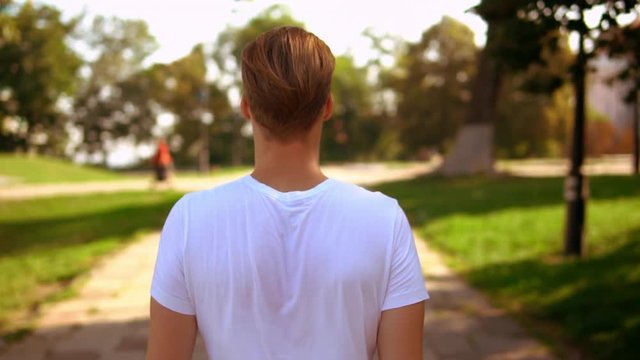 back view caucasian man with blond hair walking in park. guy wearing white t-shirt outdoors slow motion