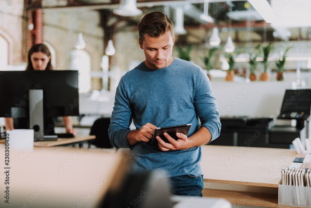 Wall mural Waist up of a young pleasant man standing in the office while using his tablet