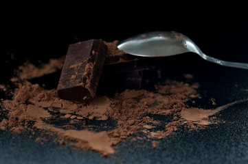 Close-up macro shot of chocolate pieces stacked on top of each other with sparse cocoa and spoon on black background. Selective focus 