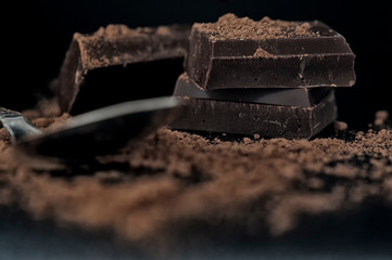 Close-up macro shot of chocolate pieces stacked on top of each other with sparse cocoa and spoon on black background. Selective focus 