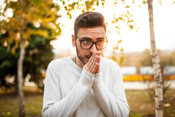 Young handsome hipster man in glasses and a white sweater is walking in a cold autumn park. Heats his hands with his breath. Concept of seasonal autumn cold