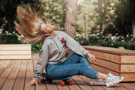 Young woman in jeans sitting on the skateboard in park. Lifestyle. Flying hairs