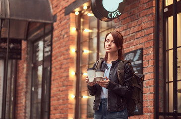 Obraz na płótnie Canvas A charming girl wearing a leather jacket with a rucksack holding cup with takeaway coffee outside near the cafe.