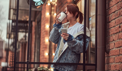 A beautiful tattooed girl wearing a denim coat drinks takeaway coffee holding a smartphone outside the cafe.