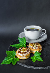 Two cake biscuits with white cream, banana slices and chocolate and a cup of tea coffee on a slate dish on a black background, decorated with green ivy leaves. Free space for text. Detailed. Vertical