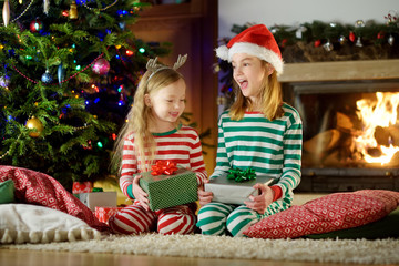 Happy little sisters wearing Christmas pajamas opening gift boxes by a fireplace in a cozy dark living room on Christmas eve.
