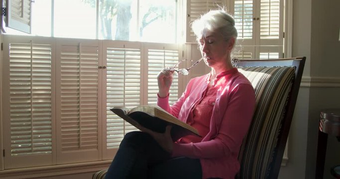 A senior adult Caucasian woman sitting by a bright window reading Bible checks her glasses before continuing.