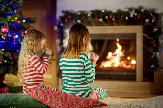 Two Cute Happy Girls Having Hot Chocolate By A Fireplace In A Cozy Dark Living Room On Christmas Eve.